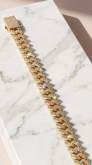 A gold-toned Cuban link bracelet covered in sparkling clear stones, displayed diagonally on a white marble rectangular block against a soft beige background.